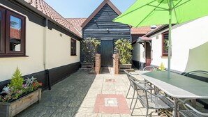 Un patio con una mesa y sillas, una sombrilla verde, y un edificio con fachada azul oscuro y techo de tejas rojas.