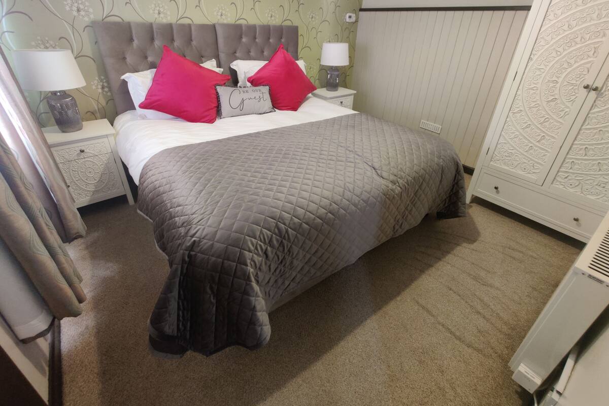 Cama bem arrumada com colcha cinza, duas almofadas vermelhas e uma almofada decorativa com texto. Há duas mesas de cabeceira com abajures, um guarda-roupa branco e piso coberto por carpete.
