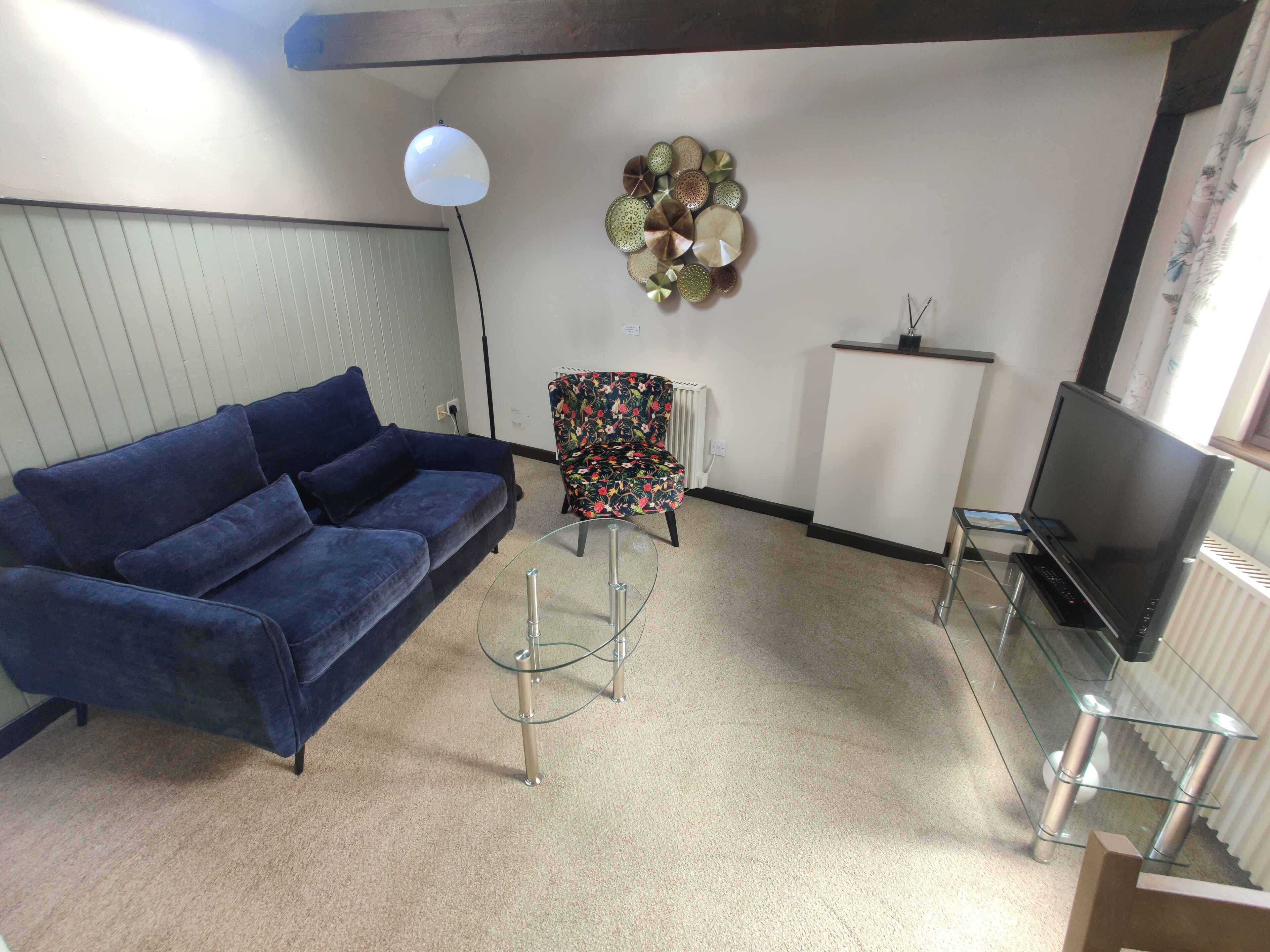 Un salon avec un canapé bleu, une table basse en verre, un fauteuil à motifs floraux, une assiette décorative accrochée au mur, un radiateur et un téléviseur posé sur un meuble.