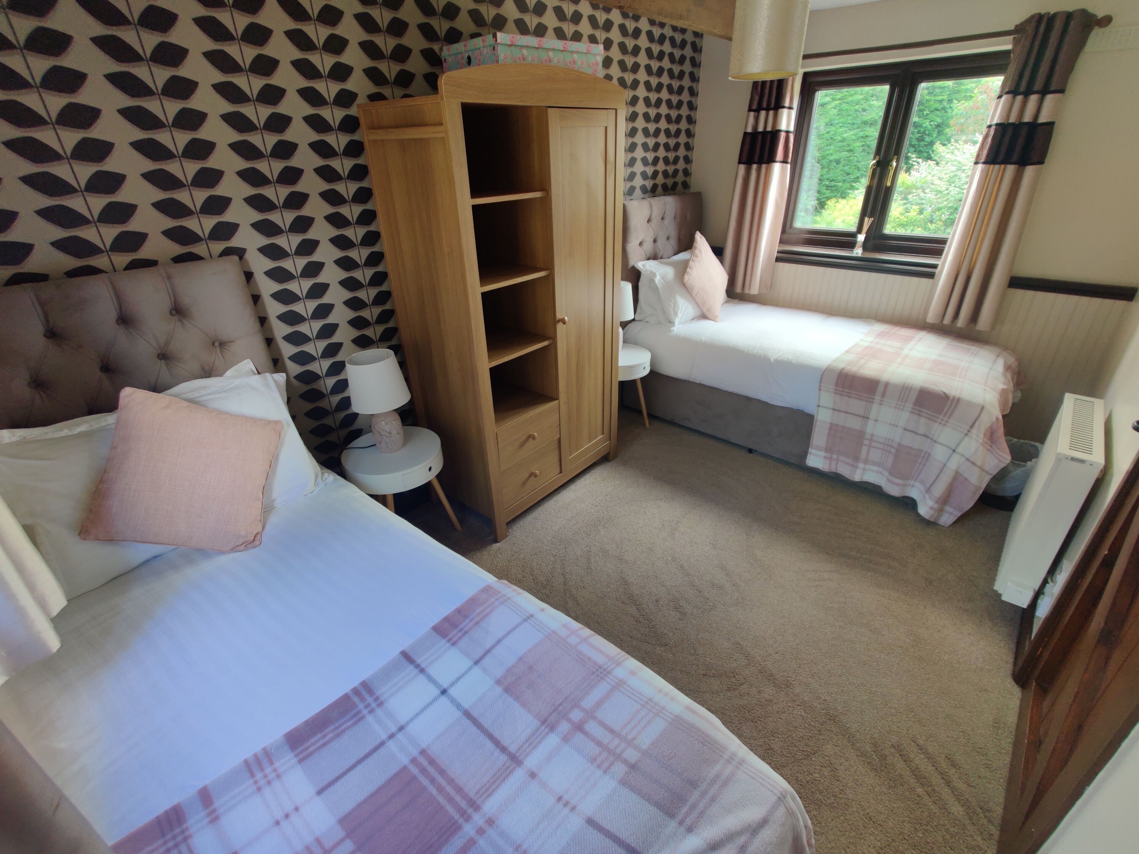 A bedroom with two beds, a wardrobe, a bedside table with a lamp, and a window with curtains.