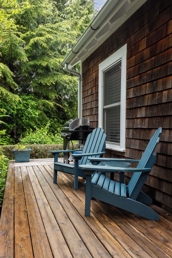 Una terraza de madera con dos sillas Adirondack y una parrilla, frente a una casa con revestimiento oscuro y una ventana con persianas.