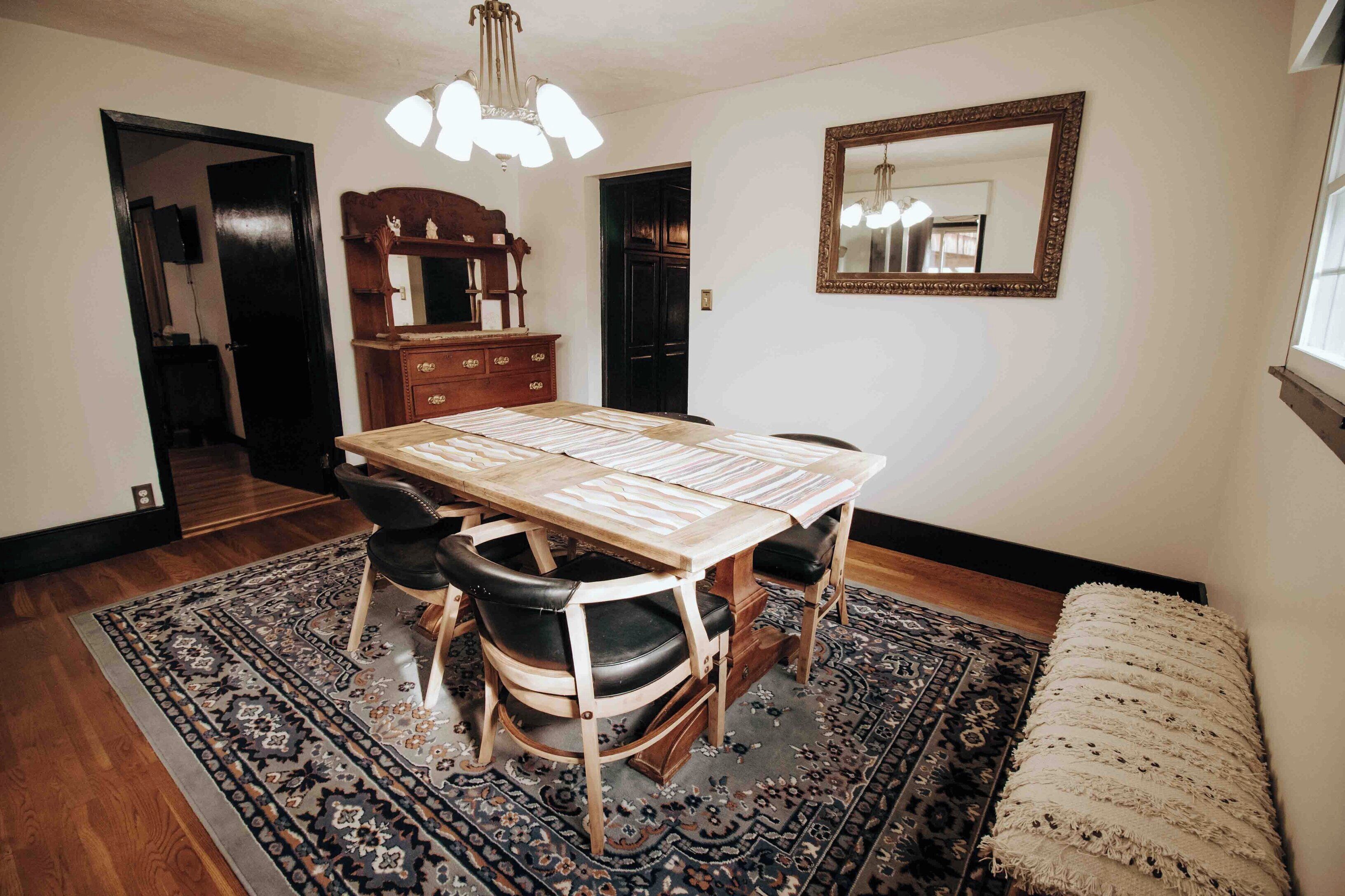 Zona pranzo con tavolo in legno, sedie in pelle nera, tappeto decorato, specchio e una panca.