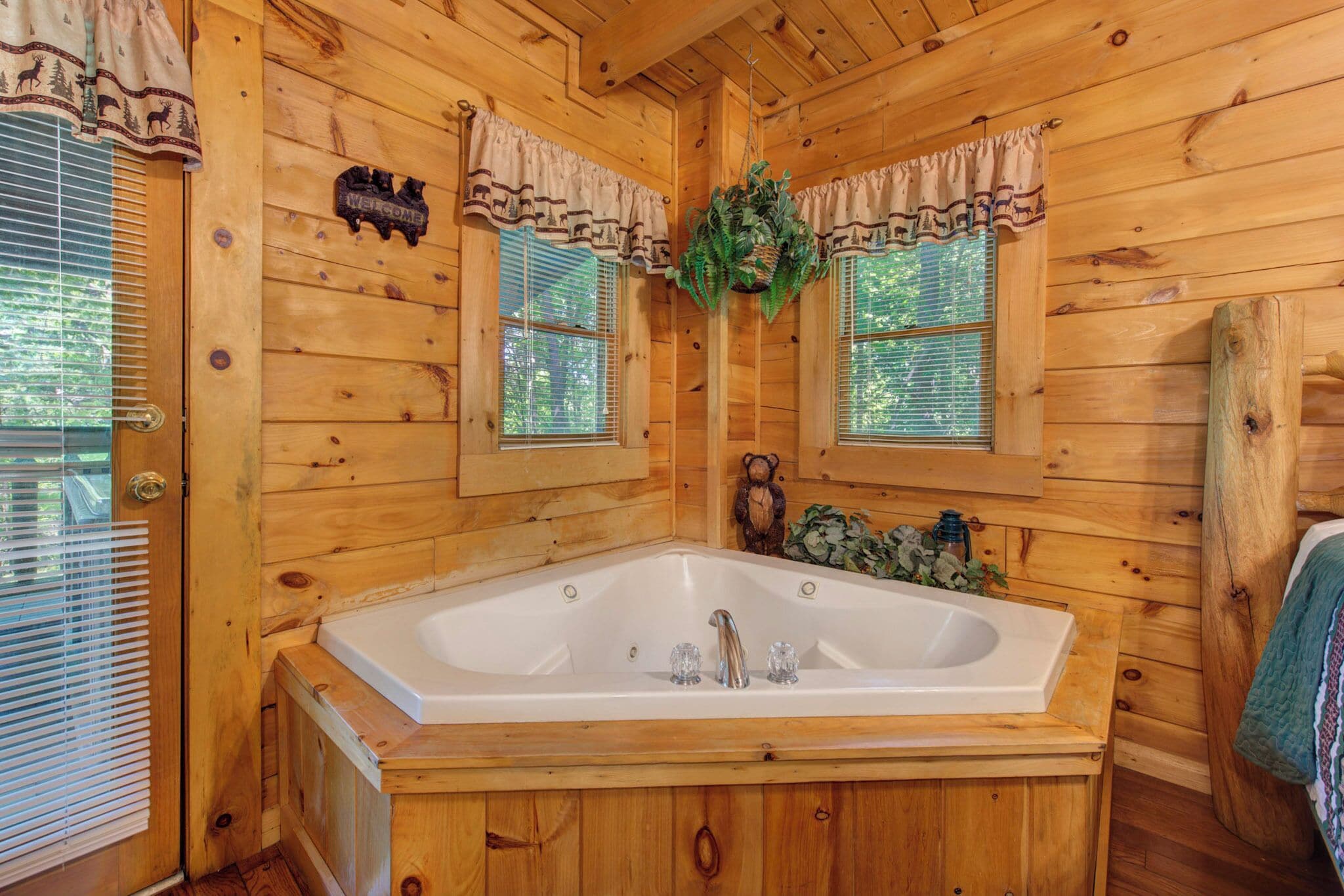 A wooden cabin bathroom with a large bathtub, two windows with curtains, and a welcome sign.