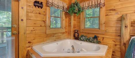 A wooden cabin bathroom with a large bathtub, two windows with curtains, and a welcome sign.