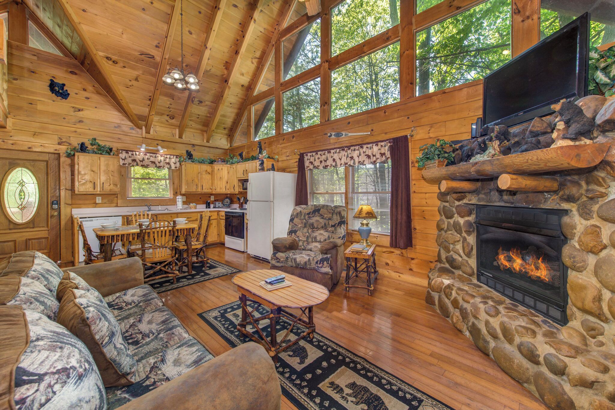 A log cabin interior with a stone fireplace, wooden furniture, a dining area, and large windows.