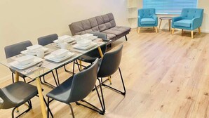 Une pièce comprenant une table à manger dressée pour six personnes, un canapé, deux fauteuils et une grande fenêtre avec des stores.