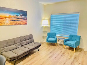 A living room with a grey sofa, two blue armchairs, a small table, a lamp, and a large window with blinds. - 2 Bd, 2.5 Baths, Sleeps 6/G9 (Reno)