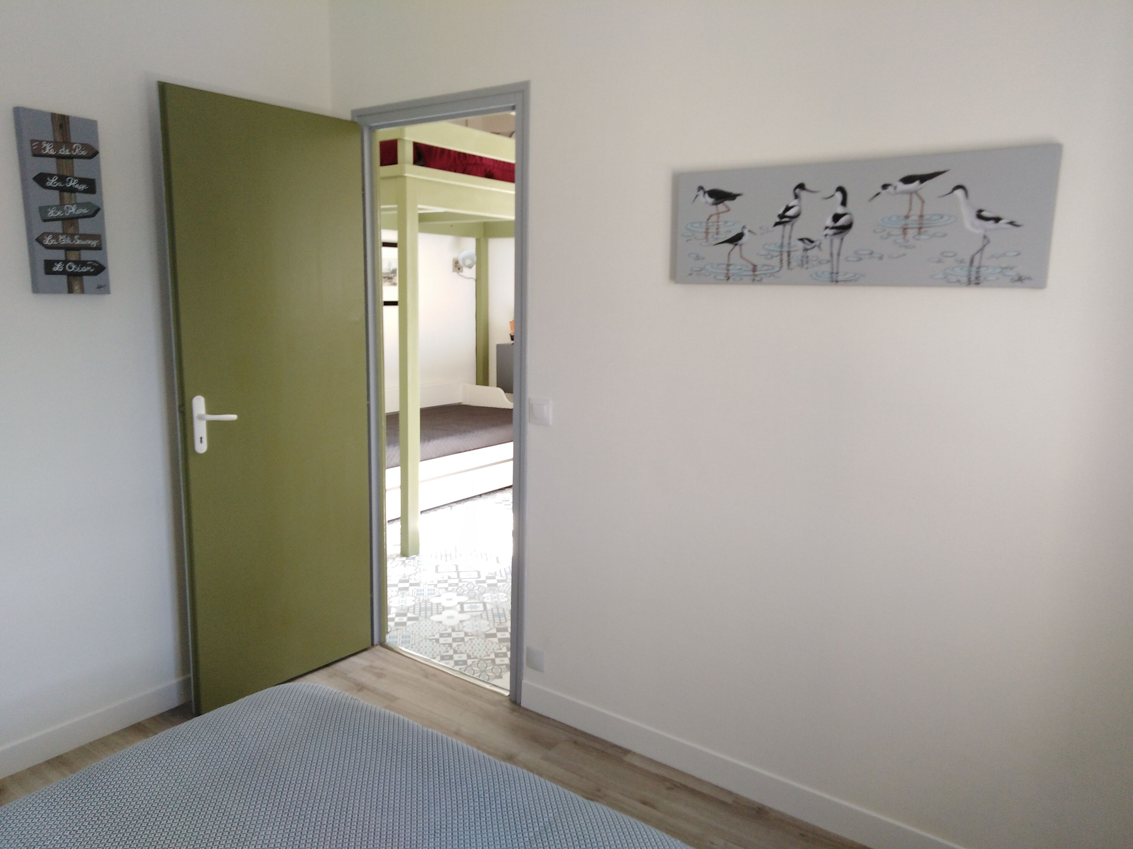 A room with a green door, a bed with a patterned bedspread, a wall with a bird painting, and a sign with text.