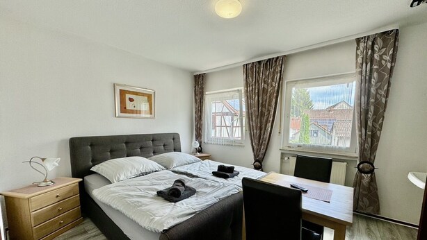 A bedroom with a bed, bedside table, chair, desk, and windows with curtains. - Gasthaus Nangfa Engel  (Edermünde)