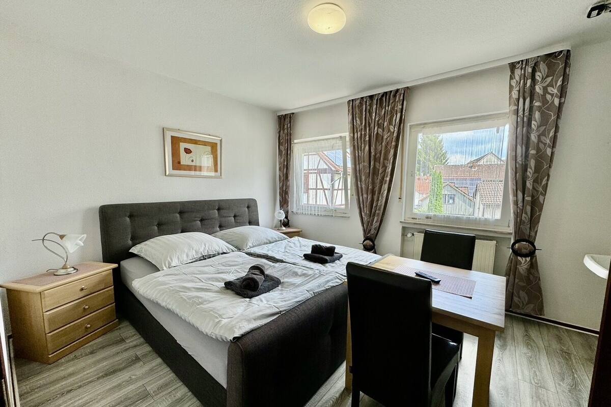 A bedroom with a bed, bedside table, chair, desk, and windows with curtains.