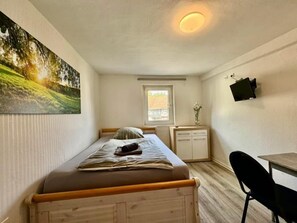 A small, single-bed room with a wooden headboard, a desk, a chair, a TV mounted on the wall, and a window with a view of a building. - Gasthaus Nangfa Engel  (Edermünde)
