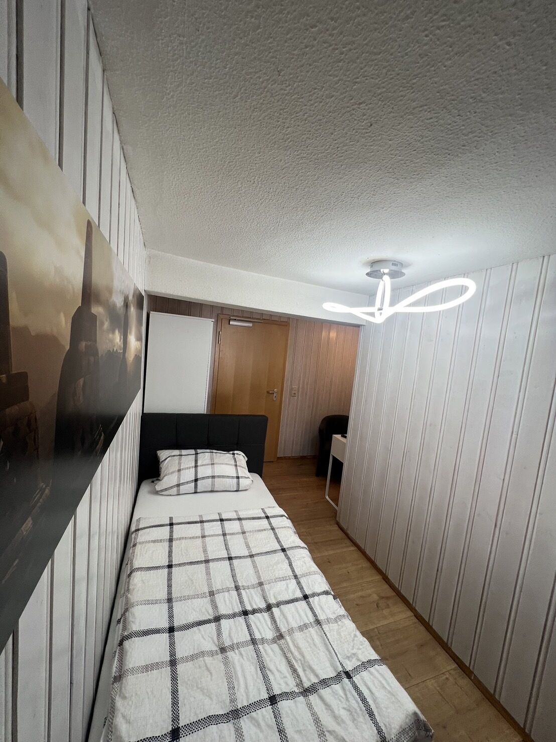 Habitació estreta amb un llit, un ventilador de sostre i una porta que dona a una altra habitació.