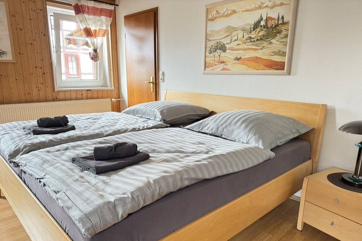 A double bed with grey bedding, a wooden headboard, and a nightstand with a lamp.