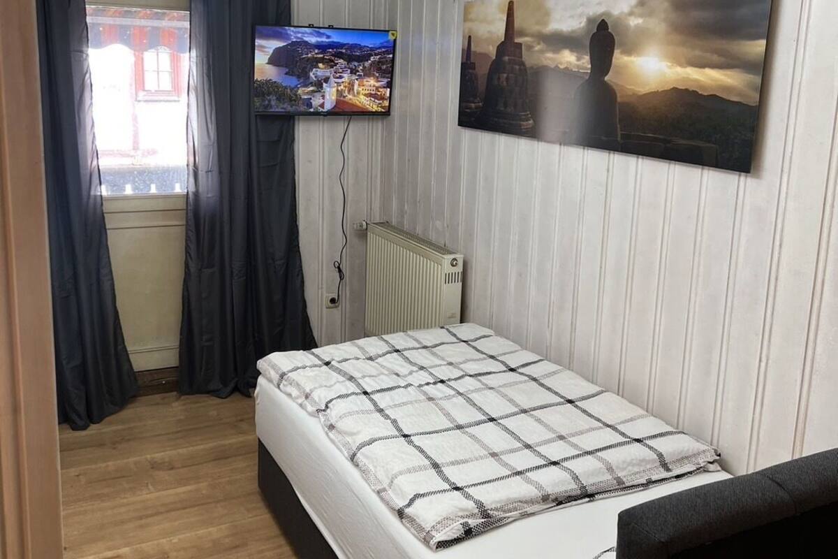 A bedroom with a bed, a TV mounted on the wall, a window with curtains, and a radiator.
