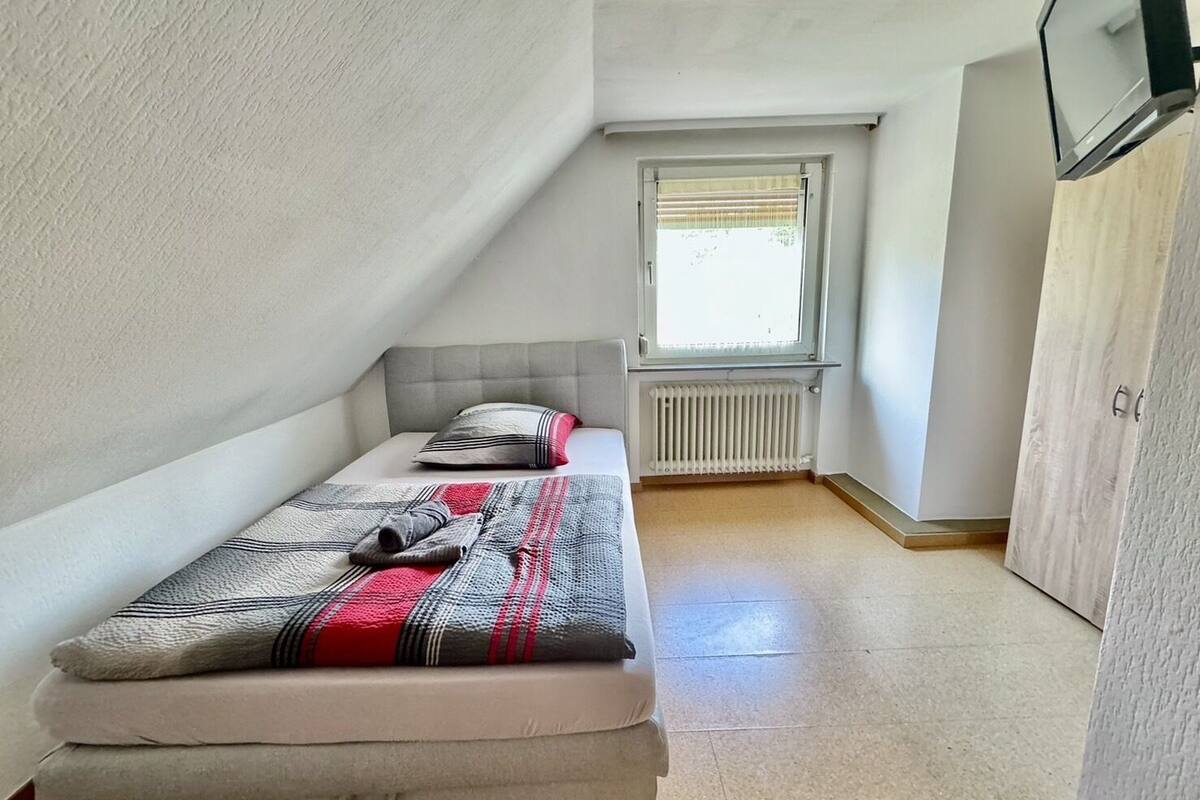 A small, single-bed room with a sloped ceiling, a radiator, a window with blinds, and a wardrobe.