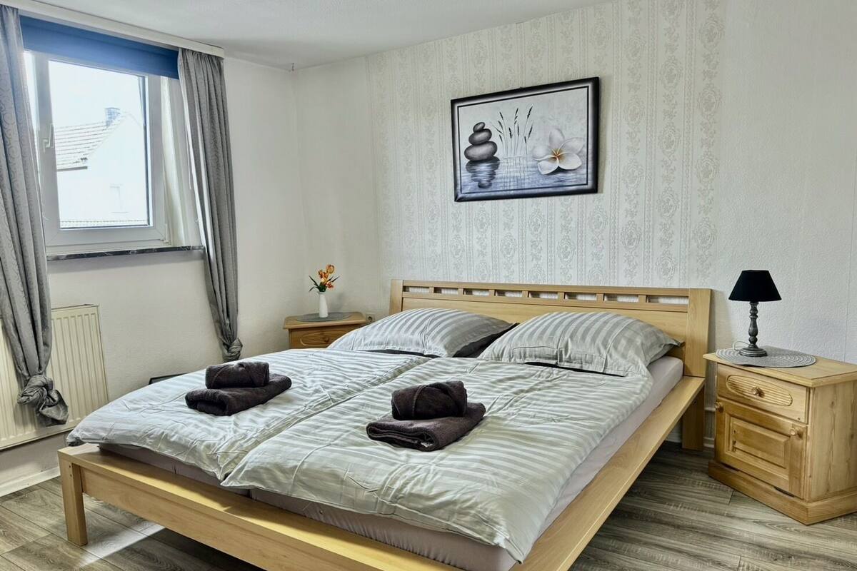 A neatly made bed with pillows and towels, a bedside table with a lamp, and a framed picture on the wall.