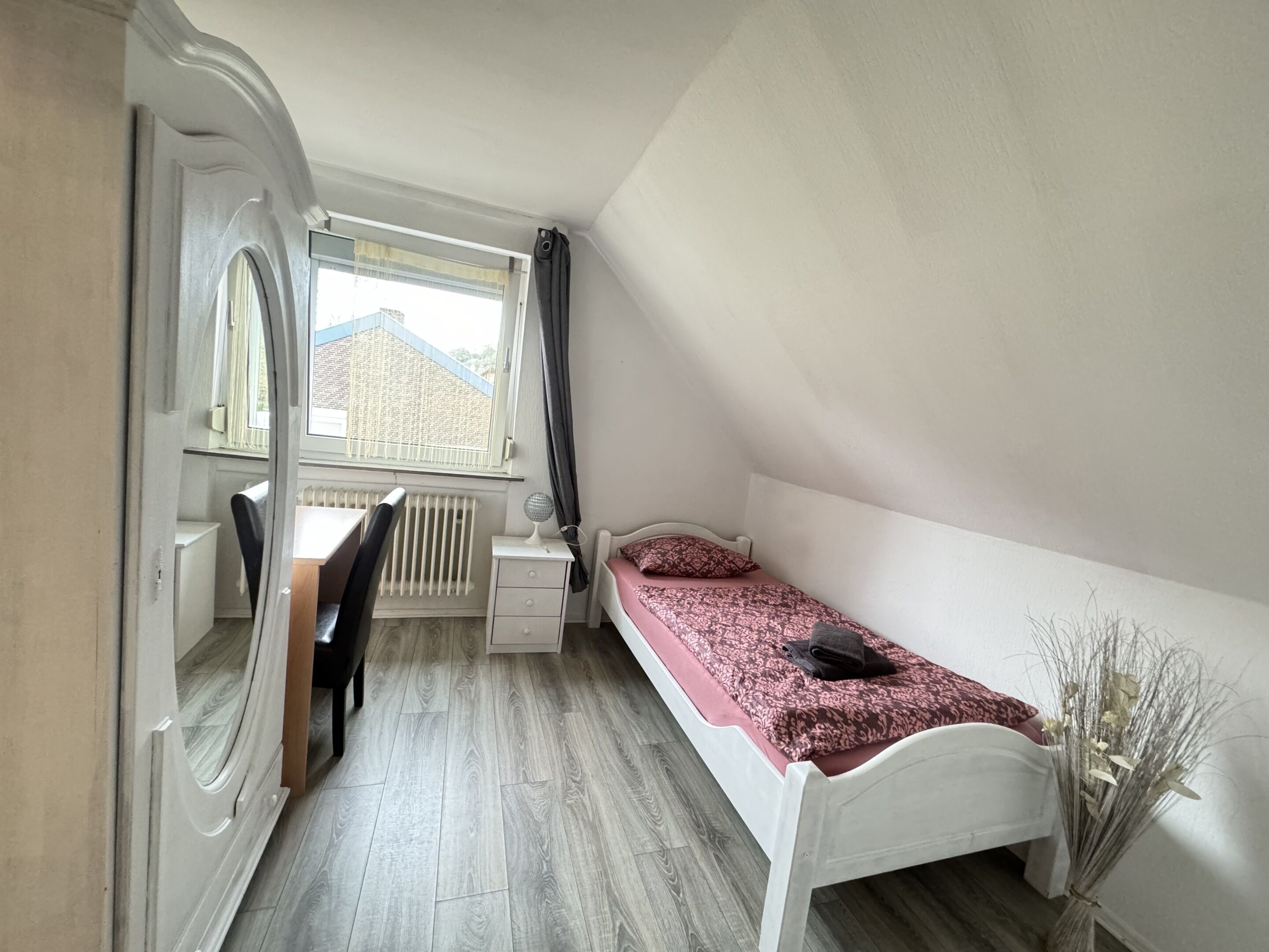 Una habitación pequeña y bien iluminada con una cama individual, un escritorio y una silla. Se aprecia una ventana con vistas a edificios.