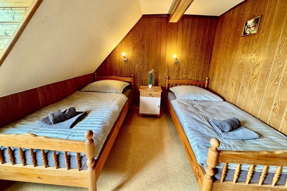A small, cozy attic bedroom with two wooden beds, a nightstand, and a wall-mounted light fixture.