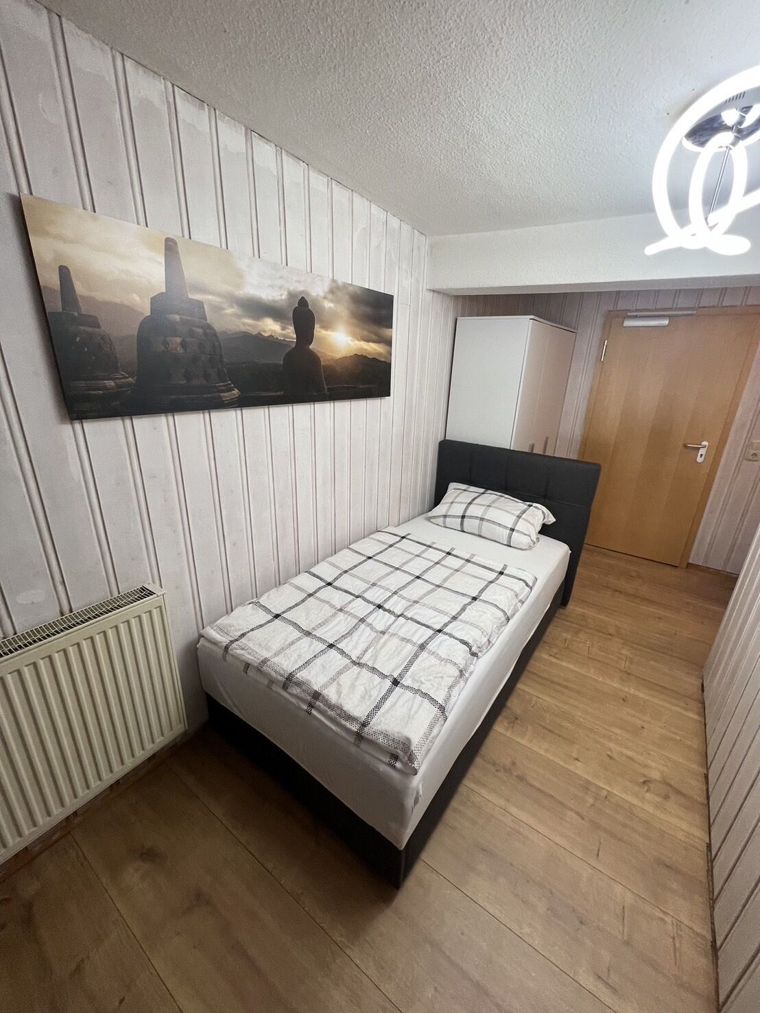 Ein Schlafzimmer mit einem Bett, einem Heizkörper, Holzboden, einer Tür und einem an der Wand hängenden Bild.