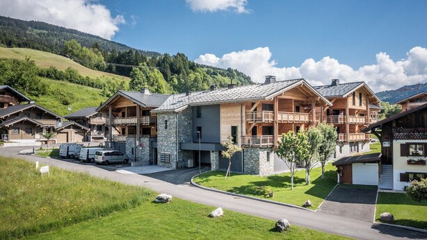 Une rangée de chalets de style rustique, construits en rondins de bois et dotés de fondations en pierre, avec une allée pavée et un paysage montagneux en toile de fond.