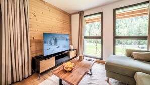 Un salon moderne avec un panneau mural en bois, un téléviseur à écran plat, une table basse en bois sur laquelle sont disposés des fruits, un canapé et de grandes fenêtres donnant sur une forêt.