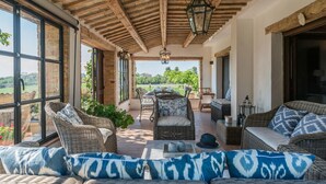 A covered patio with wicker furniture, a dining table, and large windows offering a view of the countryside.
