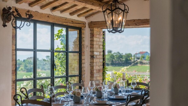 A dining area with a table set for six, large windows with a view of greenery and a distant building, wooden beams on the ceiling, and a hanging lantern.