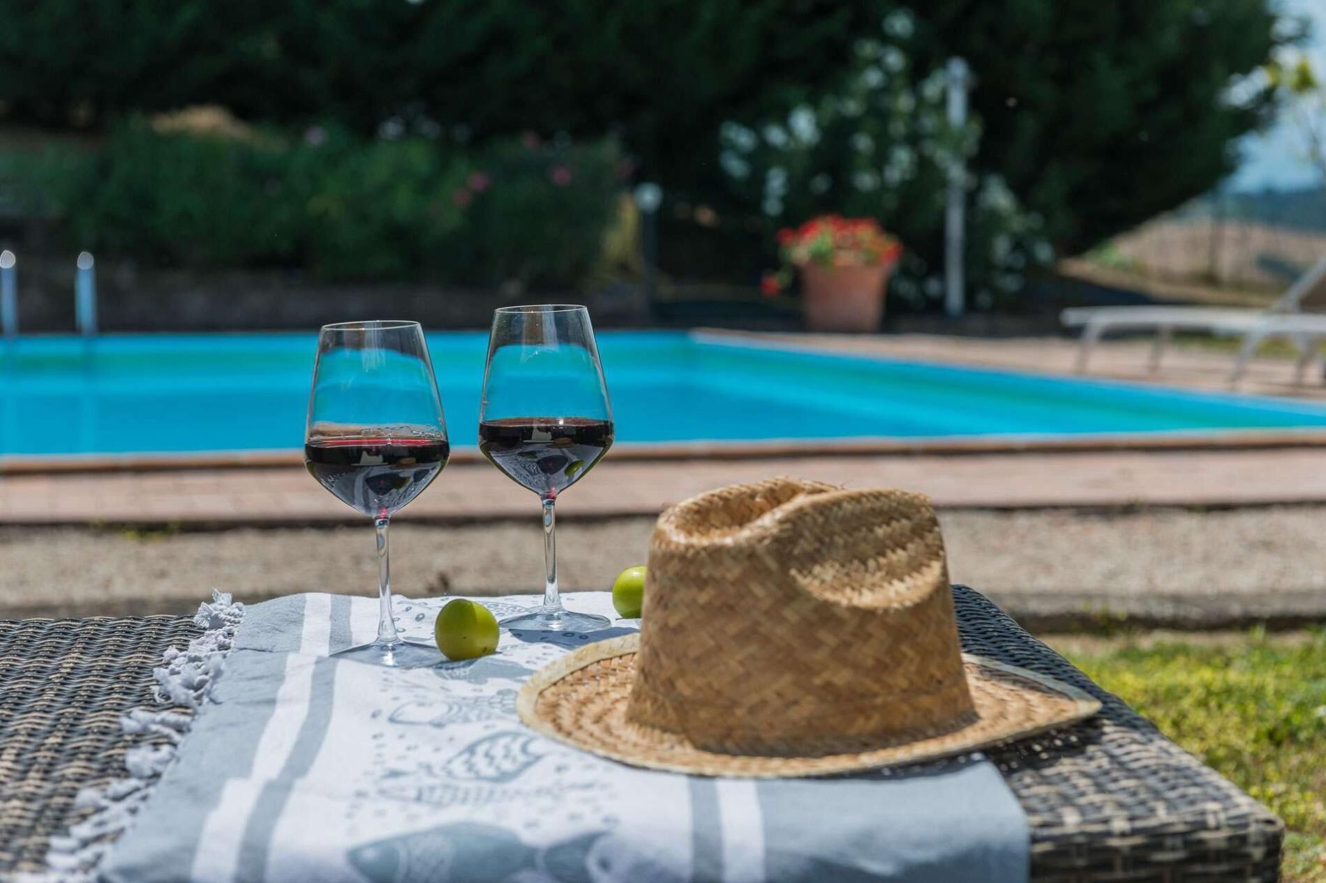 A wicker chair with a straw hat and two glasses of red wine on a patterned tablecloth by a pool.