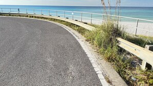 Une route sinueuse bordée d’une rambarde métallique, qui mène à une plage aux eaux d’un bleu limpide et au rivage sablonneux.