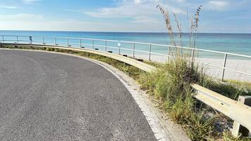 Eine gewundene Straße mit Metallgeländer, die zu einem Strand mit klarem, blauem Wasser und sandigem Ufer führt.