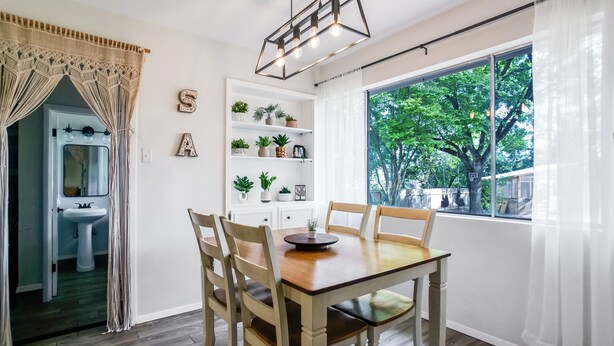 A dining area with a wooden table and chairs, a white wall with shelves holding plants, a window with a view of trees, and a door leading to a bathroom. - The TX Oasis.  Boho Paradise.  Huge Backyard. (San Antonio)