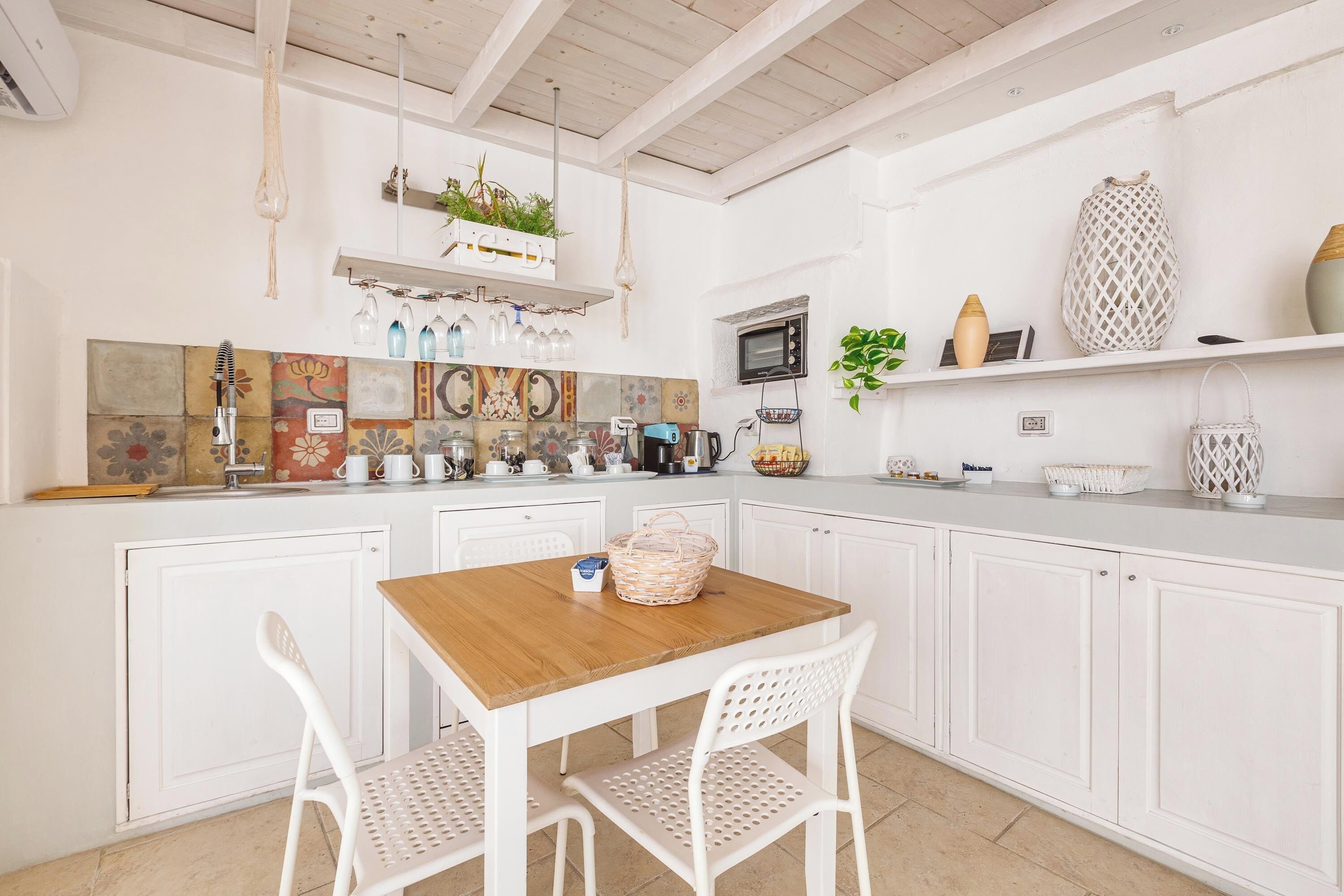 Una cucina con mobili bianchi, un tavolo in legno e sedie bianche. Si vedono un forno a microonde, un lavandino e diversi oggetti da cucina sul ripiano.