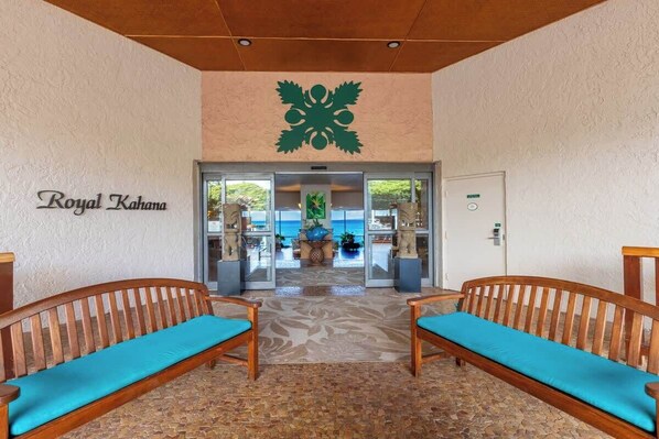A reception area with a sign reading 'Royal Kahana,' two wooden benches with blue cushions, a large green emblem on the wall, and a view of the ocean through glass doors.