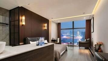 A modern hotel room with a large bed, a sitting area, a desk, a view of the city, and a bathroom.