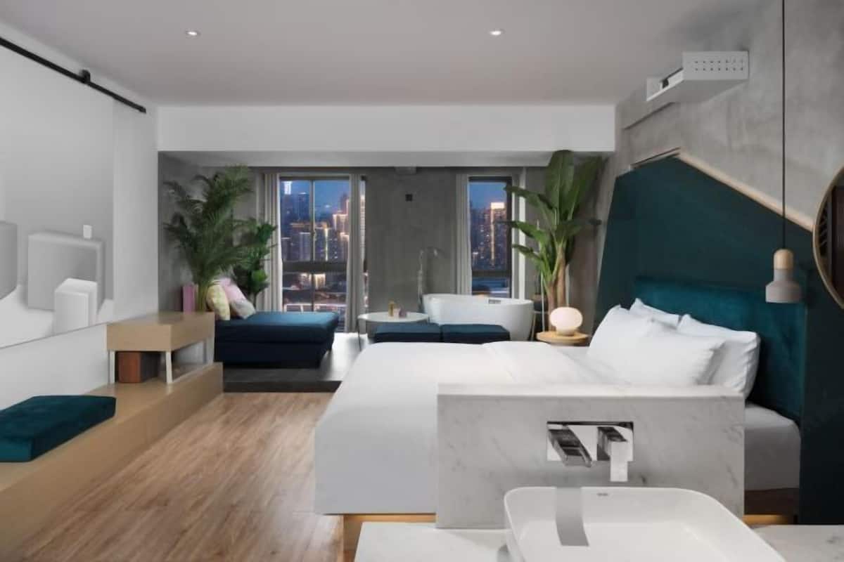 Ein modernes Hotelzimmer mit einem großen Bett, einer Sitzecke, einem kleinen Tisch und Blick auf die Stadtlandschaft.