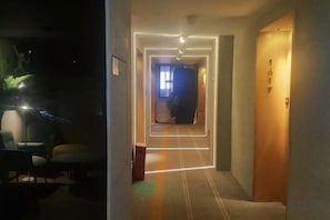 Een hotelgang met vloerbedekking, een houten deur met nummer en een lichtbron aan het einde.