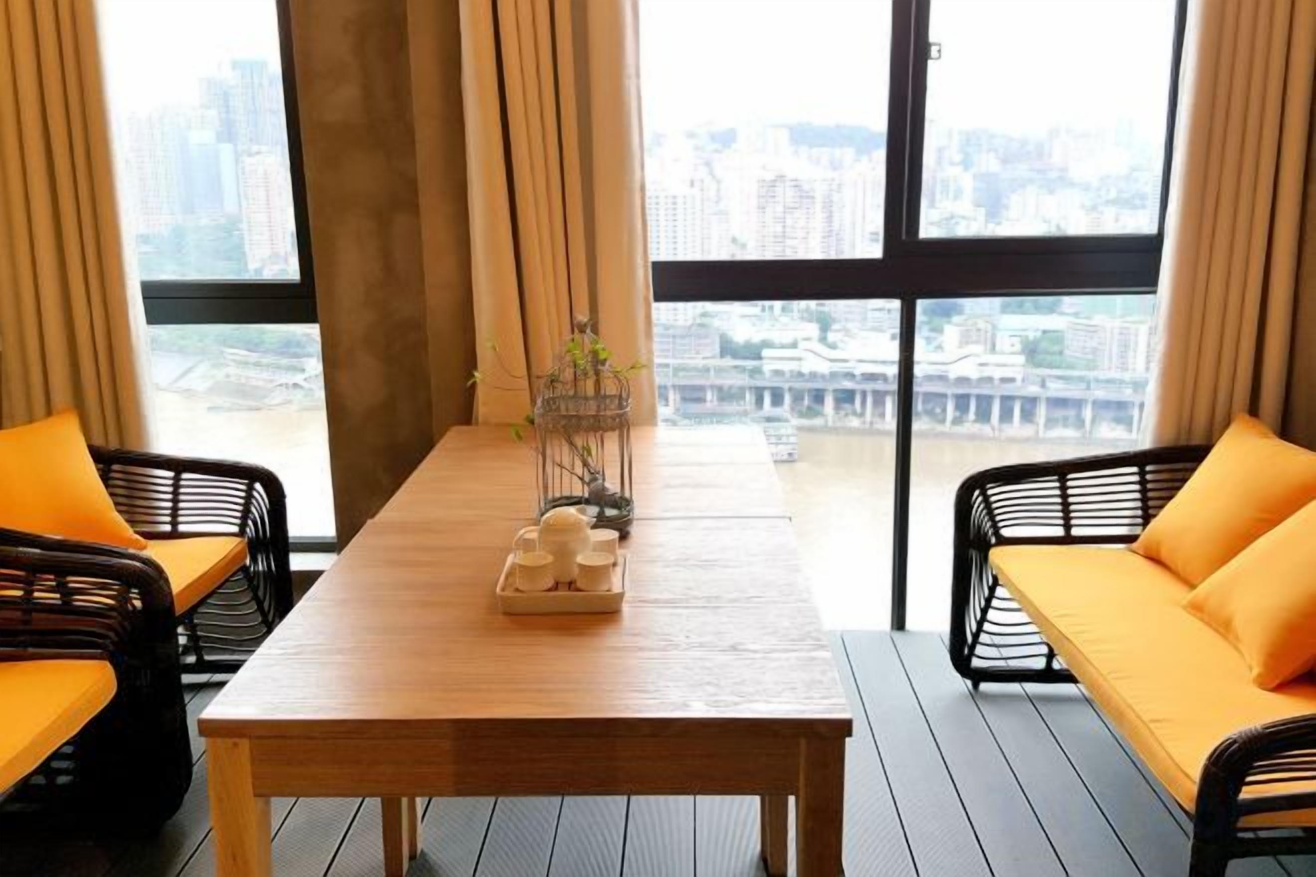 A room with a wooden table, two black wicker chairs with orange cushions, a window with a view of buildings, and a curtain.