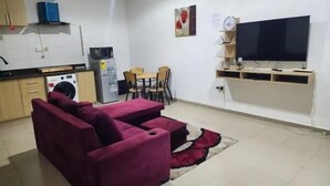 A compact living space with a sofa, a dining table with chairs, a TV, a washing machine, and a small kitchen area. - Masterpiece 1 bedroom  (Accra)