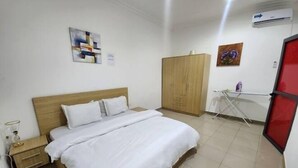 A hotel room with a bed, a wooden wardrobe, a small table with a lamp, and a red-framed window. - Masterpiece 1 bedroom  (Accra)