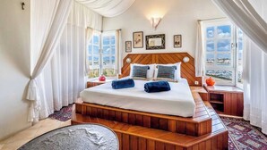 A bedroom with a wooden headboard, a large bed with white linens, two bedside tables, and a view of the city through large windows.