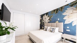 A modern bedroom with a bed, a flat-screen TV, a potted plant, and a wall with a floral mural. - Villa Oceano by Indigo (Carvoeiro)