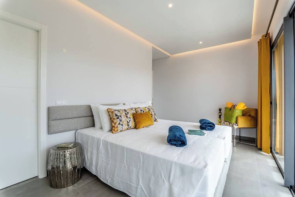 A modern bedroom with a large bed, a bedside table, a small stool, and a window with curtains.