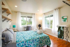 Une chambre à coucher avec un lit, un bureau, une chaise, un ventilateur et une fenêtre avec des rideaux.