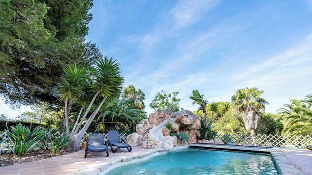 A swimming pool with a waterfall feature, surrounded by palm trees and a stone wall. - Villa Concha by Indigo (Carvoeiro)