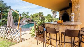 An outdoor bar area with a stone fireplace, a pool, and a view of a tropical landscape. - Villa Concha by Indigo (Carvoeiro)