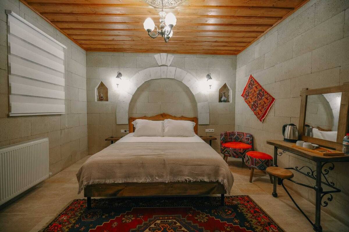 A bedroom with a wooden ceiling, a bed with a headboard, a nightstand, a chair, a small table with a mirror, and a radiator.