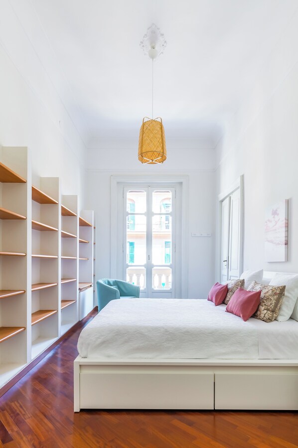 Een slaapkamer met een bed, witte muren, houten vloer, een deur met ramen en een hangende lamp.