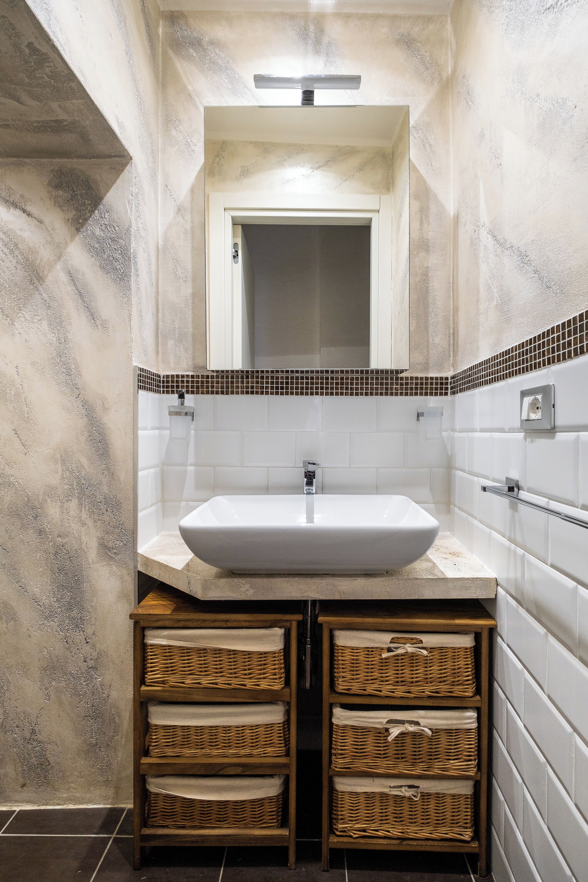 Un baño con un lavamanos blanco, un espejo encima y un reverso empotrado de azulejos. Hay cestos de mimbre debajo del lavamanos.