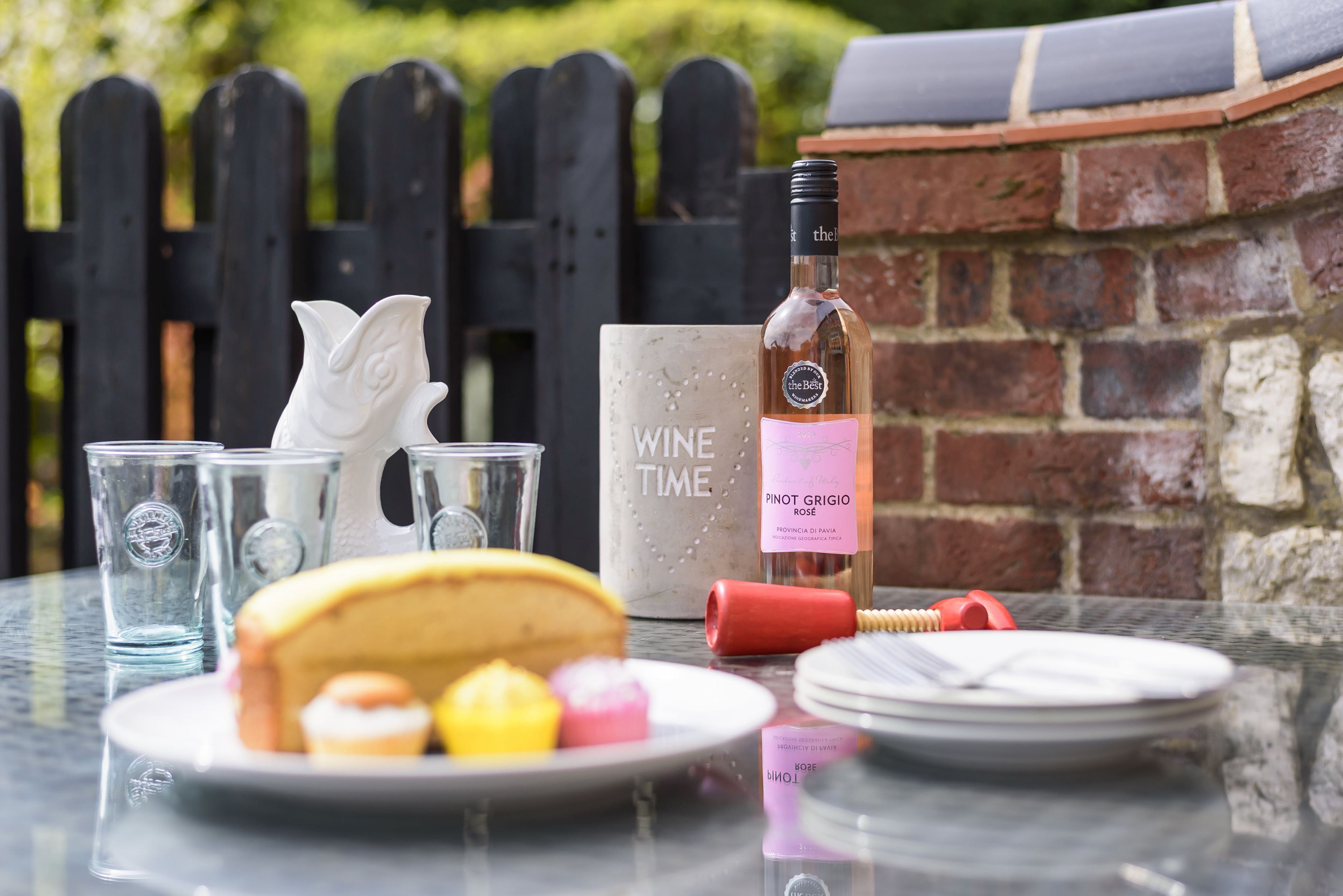 A table with a bottle of Pinot Grigio Rosé, a cake, and a decorative item labeled "WINE TIME" in a garden setting with a brick wall and wooden fence in the background.