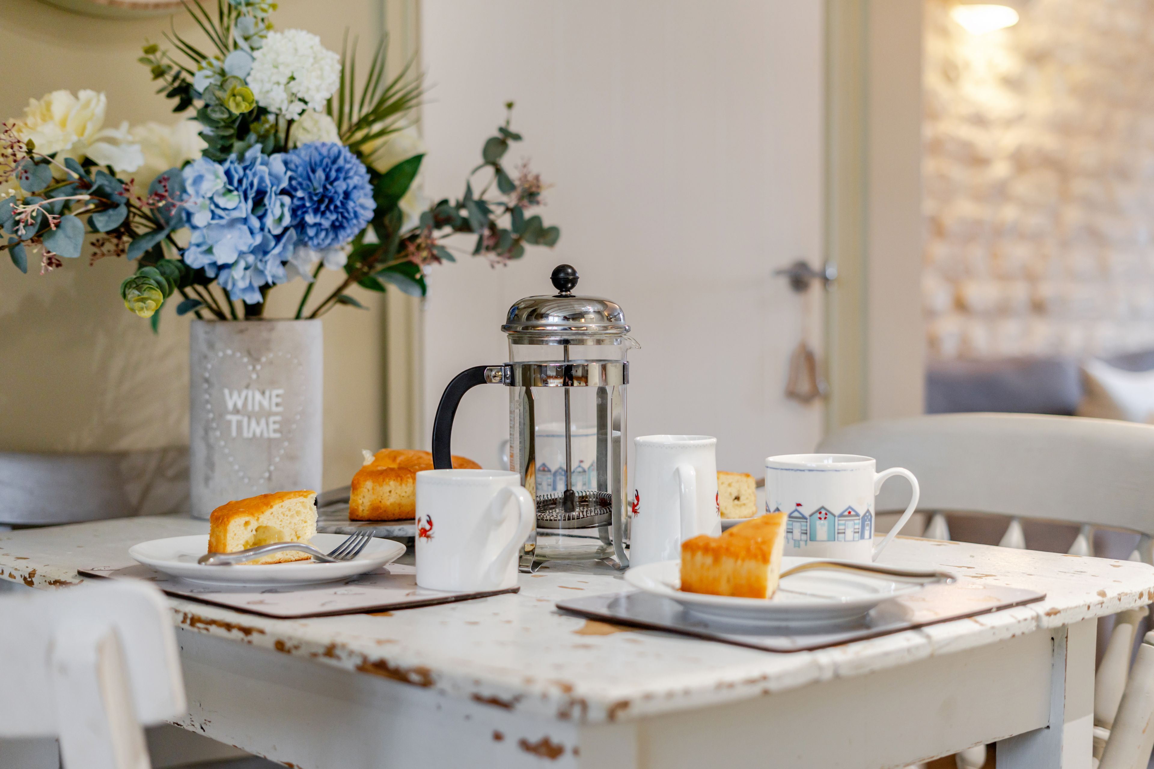 Une table rustique agrémentée d’une cafetière, de tasses blanches et d’un arrangement floral.